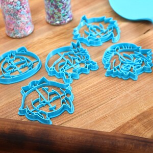 Pu&ograve; includere: Cinque tagliabiscotti in plastica blu a forma di facce di cartoni animati su una superficie di legno. I tagliabiscotti sono disposti a semicerchio.
