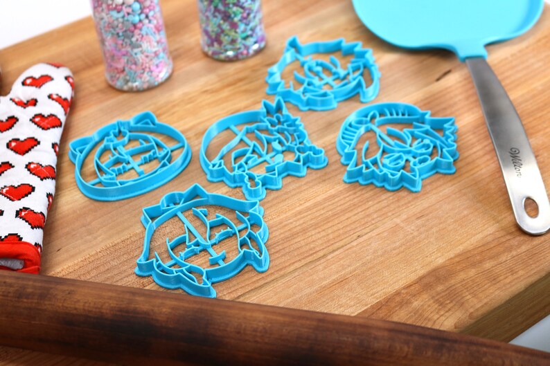 Pu&ograve; includere: Cinque tagliabiscotti in plastica blu a forma di facce di cartoni animati su una superficie di legno. I tagliabiscotti sono disposti a semicerchio.