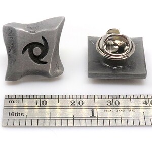 May include: A silver metal pin with a black swirl design. The pin is about 1 inch in diameter and has a safety clasp on the back.