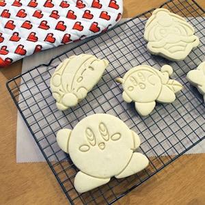 Pode incluir: Cinco biscoitos brancos de a&ccedil;&uacute;car em uma grelha de resfriamento de arame. Os biscoitos t&ecirc;m a forma de personagens de desenhos animados.