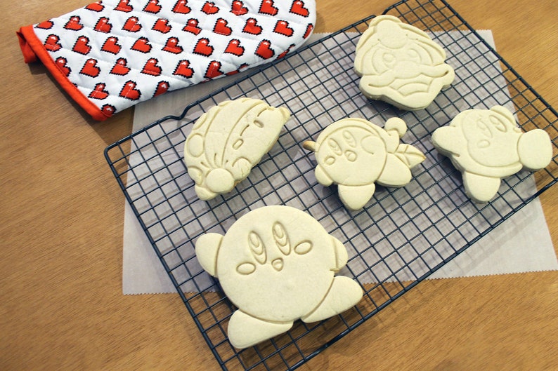Pode incluir: Cinco biscoitos brancos de a&ccedil;&uacute;car em uma grelha de resfriamento de arame. Os biscoitos t&ecirc;m a forma de personagens de desenhos animados.