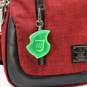 May include: A red and black crossbody bag with a green shield-shaped charm hanging from the zip. The charm has a white symbol inside. The bag has a black leather trim and silver zips.