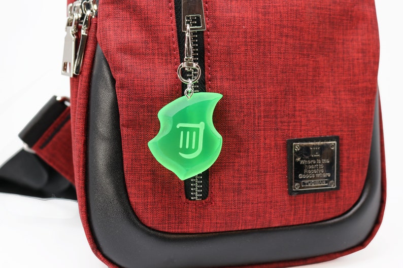 May include: A red and black crossbody bag with a green shield-shaped charm hanging from the zip. The charm has a white symbol inside. The bag has a black leather trim and silver zips.