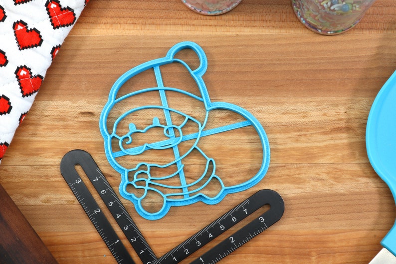 Op de afbeelding: Een blauwe plastic koekjesvormer in de vorm van een cartoon kerstman die een cadeau vasthoudt. De vormer heeft een rasterpatroon aan de binnenkant.