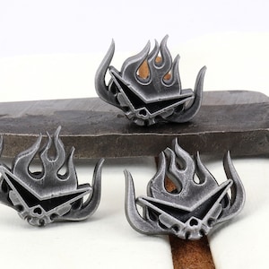 May include: Three silver metal pins with a stylized flame design and a skull and crossbones motif. The pins are on a gray metal surface.