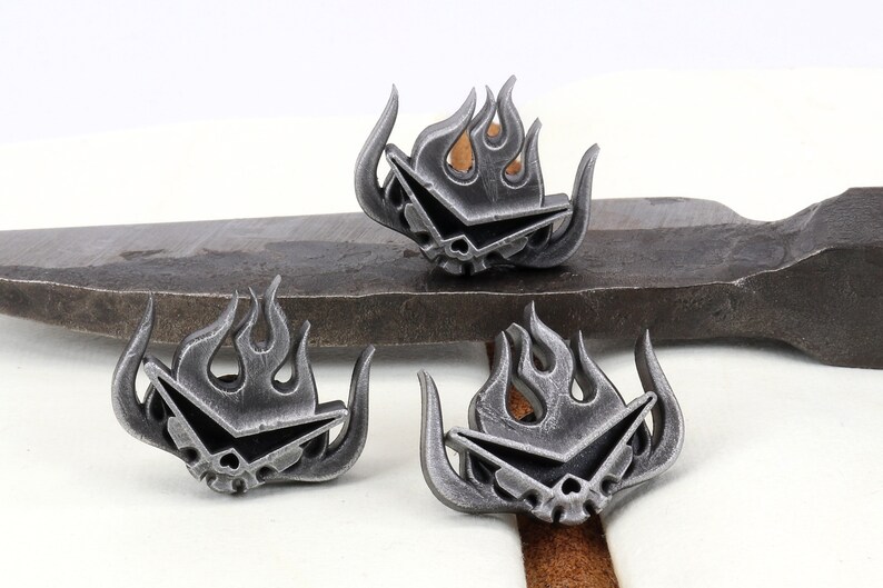 May include: Three silver metal pins with a stylized flame design and a skull and crossbones motif. The pins are on a gray metal surface.