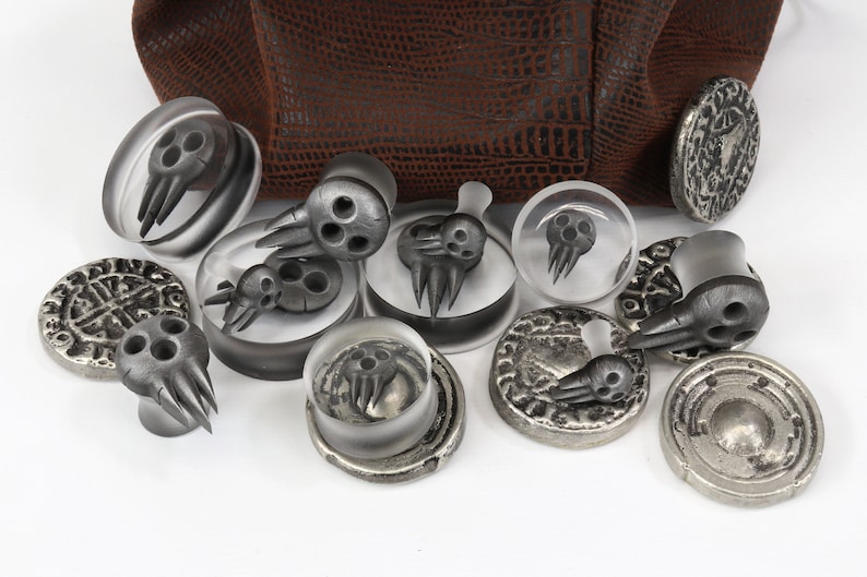 May include: A collection of gray, skull-shaped plugs and decorative metal discs. Some plugs are clear with a skull design inside. The metal discs have etched patterns. A brown textured surface is in the background.