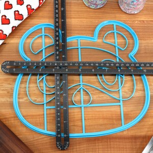 May include: A blue plastic cookie cutter shaped like an animal face with a grid pattern inside. The cutter is being measured with a black ruler.