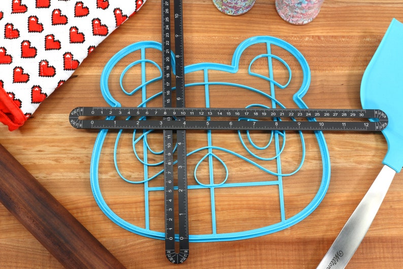 May include: A blue plastic cookie cutter shaped like an animal face with a grid pattern inside. The cutter is being measured with a black ruler.
