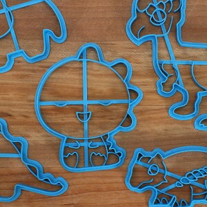 May include: A set of five blue plastic dinosaur cookie cutters. The cutters are shaped like different dinosaurs, including a triceratops, a stegosaurus, and a tyrannosaurus rex.