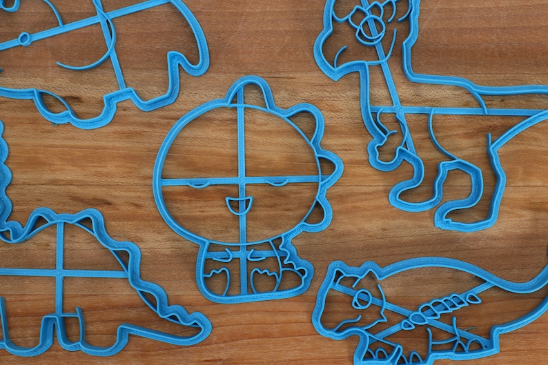 May include: A set of five blue plastic dinosaur cookie cutters. The cutters are shaped like different dinosaurs, including a triceratops, a stegosaurus, and a tyrannosaurus rex.