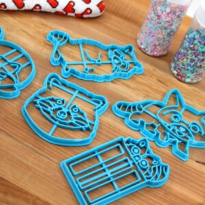Puede incluir: Un juego de seis cortadores de galletas de pl&aacute;stico azul con forma de mapaches. Los cortadores est&aacute;n sobre una superficie de madera con dos frascos de chispas en el fondo.