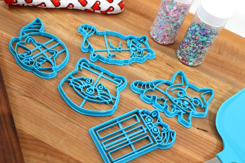 Puede incluir: Un juego de seis cortadores de galletas de pl&aacute;stico azul con forma de mapaches. Los cortadores est&aacute;n sobre una superficie de madera con dos frascos de chispas en el fondo.