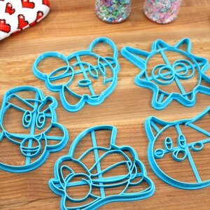 May include: Five blue plastic cookie cutters on a wooden surface. The cutters are shaped like cartoon characters, including a cat, a sun, a mouse, and two other characters.