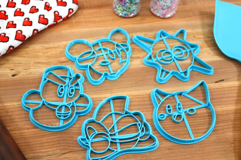 May include: Five blue plastic cookie cutters on a wooden surface. The cutters are shaped like cartoon characters, including a cat, a sun, a mouse, and two other characters.