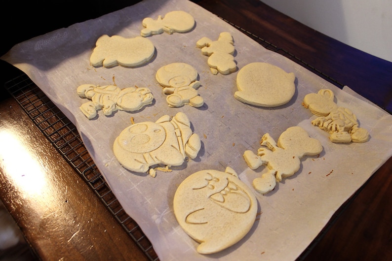May include: A sheet of parchment paper with nine unbaked sugar cookies in various shapes, including a ghost, a character with a hat, a round cookie, and a character with a long nose.