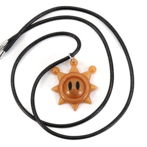 May include: A black cord necklace with a gold sun pendant. The sun has a face with two black eyes and a glittery finish.