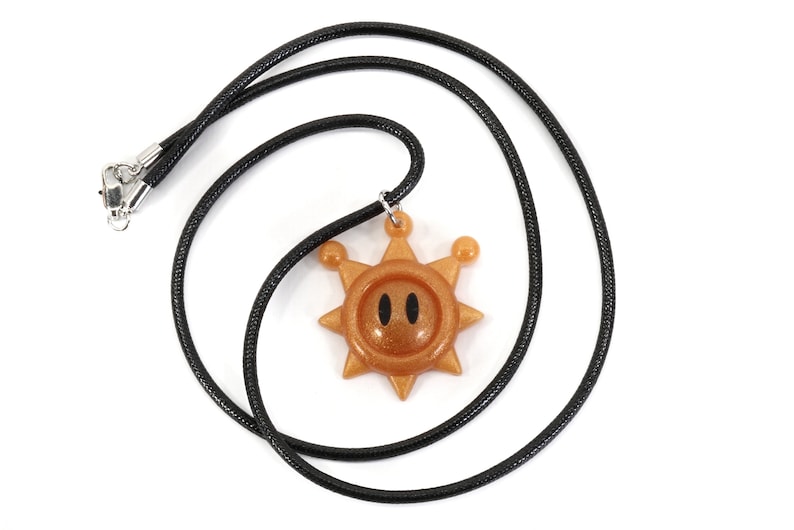 May include: A black cord necklace with a gold sun pendant. The sun has a face with two black eyes and a glittery finish.