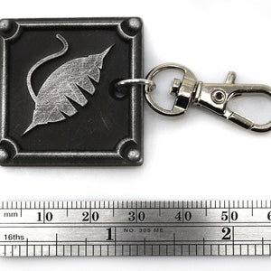 Puede incluir: Un llavero de metal plateado con un dije cuadrado negro que presenta un dise&ntilde;o de hoja estilizado. El dije tiene un anillo de metal plateado unido a &eacute;l, y el llavero tiene un broche de langosta de metal plateado.