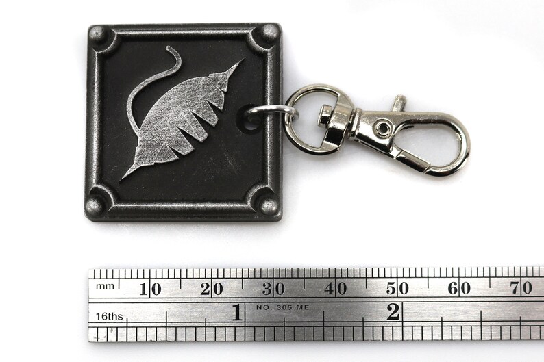 Puede incluir: Un llavero de metal plateado con un dije cuadrado negro que presenta un dise&ntilde;o de hoja estilizado. El dije tiene un anillo de metal plateado unido a &eacute;l, y el llavero tiene un broche de langosta de metal plateado.