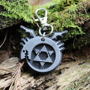 May include: A silver metal keychain featuring a circular design with a black and white symbol. The symbol is a circle with a five-pointed star inside, and a snake with fangs biting its tail.