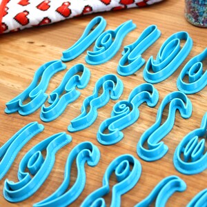 May include: A set of blue plastic cookie cutters shaped like letters of the alphabet. The cutters are arranged on a wooden surface.