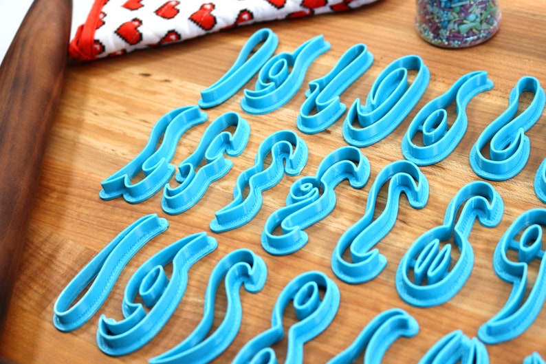 May include: A set of blue plastic cookie cutters shaped like letters of the alphabet. The cutters are arranged on a wooden surface.