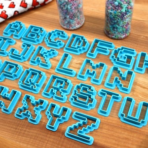 May include: A set of 26 blue plastic cookie cutters shaped like pixelated letters of the alphabet. The cutters are arranged in alphabetical order on a wooden surface.