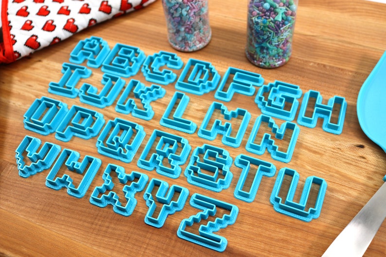 May include: A set of 26 blue plastic cookie cutters shaped like pixelated letters of the alphabet. The cutters are arranged in alphabetical order on a wooden surface.