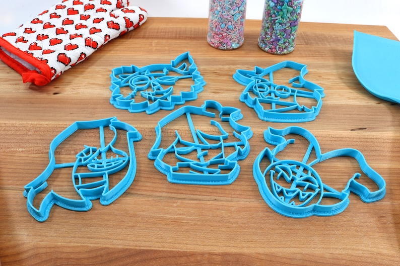 May include: Set of five blue plastic cookie cutters shaped like cartoon characters. The characters include a pirate, a monster, a ghost, a mermaid, and a kraken.