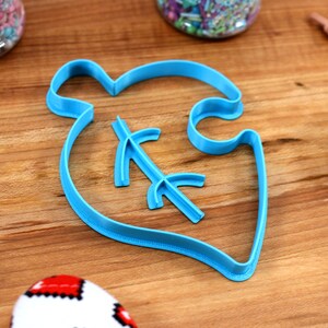 May include: A blue plastic cookie cutter shaped like a leaf with a small branch inside. The cutter is on a wooden surface.