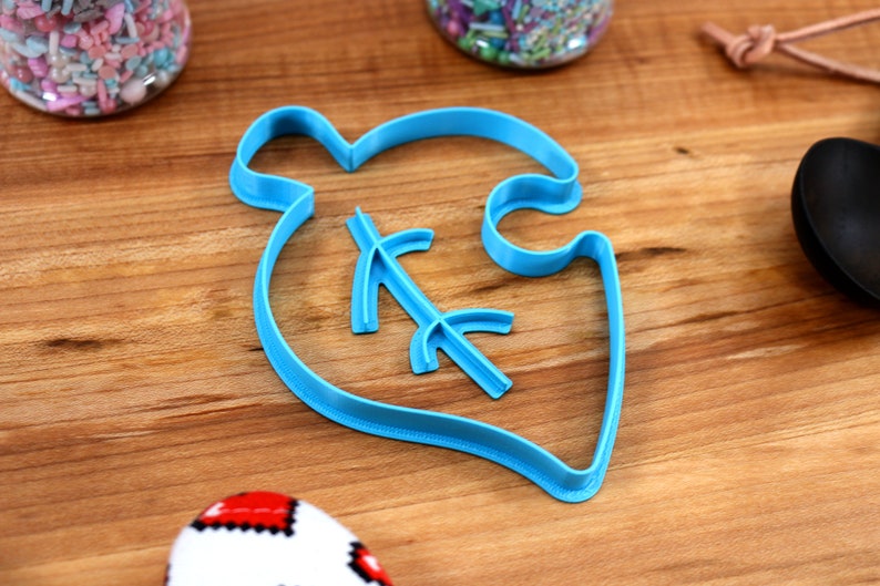 May include: A blue plastic cookie cutter shaped like a leaf with a small branch inside. The cutter is on a wooden surface.