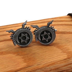 May include: A pair of silver cufflinks featuring a stylized black dragon with a star inside a circle. The cufflinks are on a wooden surface.