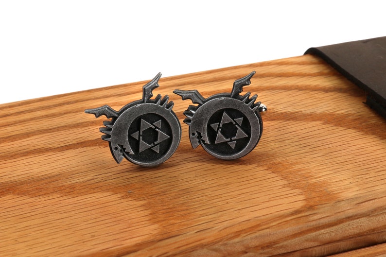 May include: A pair of silver cufflinks featuring a stylized black dragon with a star inside a circle. The cufflinks are on a wooden surface.
