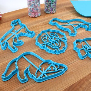 May include: Set of five blue plastic dog-shaped cookie cutters on a wooden surface. The cutters include a dog standing, a dog sitting, and three different dog head shapes.
