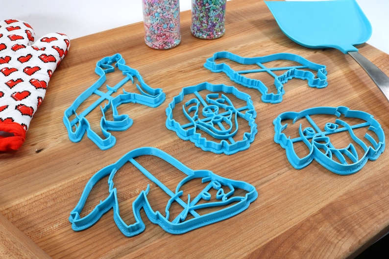 May include: Set of five blue plastic dog-shaped cookie cutters on a wooden surface. The cutters include a dog standing, a dog sitting, and three different dog head shapes.