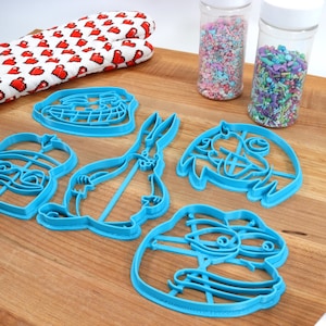 Puede incluir: Un juego de seis cortadores de galletas de pl&aacute;stico azul con forma de personajes de dibujos animados. Los cortadores est&aacute;n sobre una superficie de madera con dos frascos de chispas en el fondo.