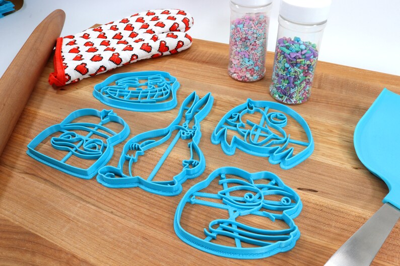 Puede incluir: Un juego de seis cortadores de galletas de pl&aacute;stico azul con forma de personajes de dibujos animados. Los cortadores est&aacute;n sobre una superficie de madera con dos frascos de chispas en el fondo.