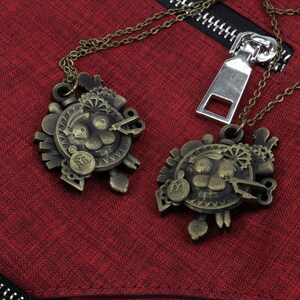 May include: Two gold-toned necklaces with intricate, detailed pendants featuring a stylized face with gears and other mechanical elements. The necklaces are hanging on a red fabric background.