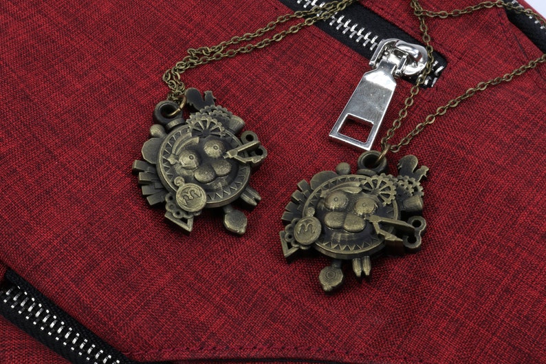May include: Two gold-toned necklaces with intricate, detailed pendants featuring a stylized face with gears and other mechanical elements. The necklaces are hanging on a red fabric background.
