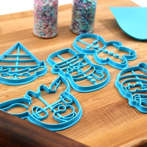 Op de afbeelding: Een set van vijf blauwe plastic kerstkoekjesvormen op een houten snijplank. De vormen zijn in de vorm van een sneeuwpop, een peperkoekman, een rendier, een zeilboot en de kerstman.