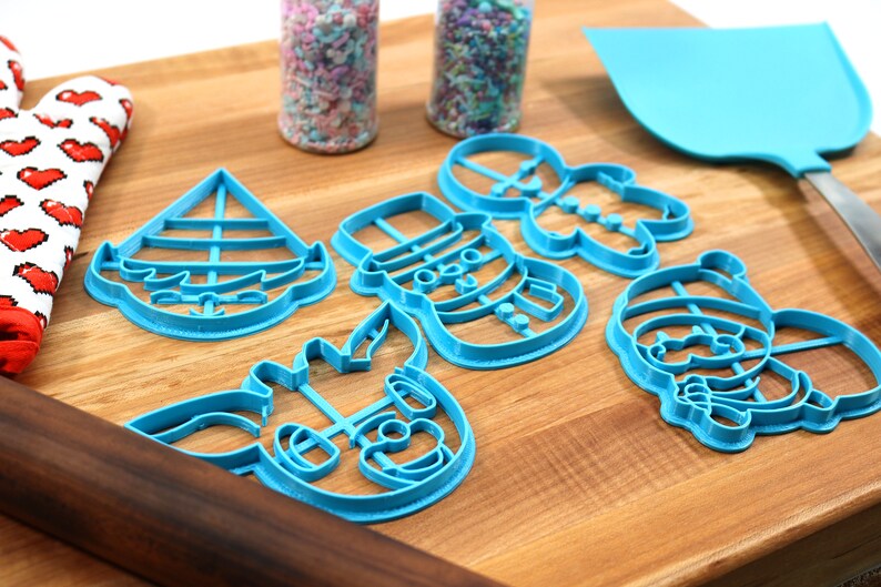 Op de afbeelding: Een set van vijf blauwe plastic kerstkoekjesvormen op een houten snijplank. De vormen zijn in de vorm van een sneeuwpop, een peperkoekman, een rendier, een zeilboot en de kerstman.