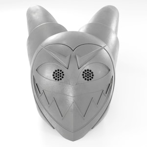 May include: A silver-colored helmet with a menacing expression, featuring two large, round eyes with black mesh details and a jagged, toothy grin.