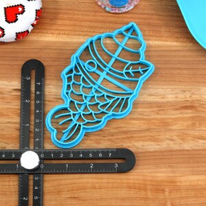 May include: A light blue plastic cookie cutter in the shape of a fish, with a detailed design. A black measuring tool is positioned near the cutter. Other items include a white and red heart-patterned item and a blue tray.