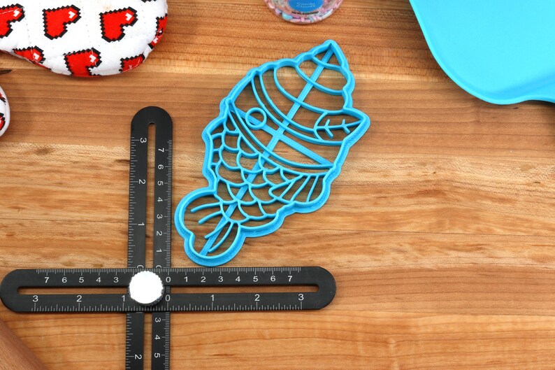 May include: A light blue plastic cookie cutter in the shape of a fish, with a detailed design. A black measuring tool is positioned near the cutter. Other items include a white and red heart-patterned item and a blue tray.