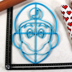 May include: A blue plastic cookie cutter in the shape of a cartoon character's face, with a large mustache and a hat with the letter "L". A wooden rolling pin and a heart-patterned oven mitt are also visible.