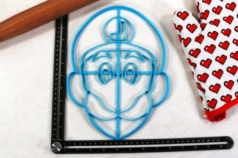May include: A blue plastic cookie cutter in the shape of a cartoon character's face, with a large mustache and a hat with the letter "L". A wooden rolling pin and a heart-patterned oven mitt are also visible.