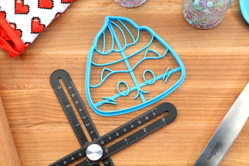 May include: A blue plastic cookie cutter shaped like a cartoon poo emoji wearing a hat. The cutter is on a wooden surface.
