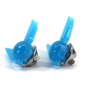 Peut inclure: Deux dos de broche en plastique bleu clair avec des fermoirs en m&eacute;tal argent&eacute;. Les dos de broche sont en forme d'aile stylis&eacute;e.