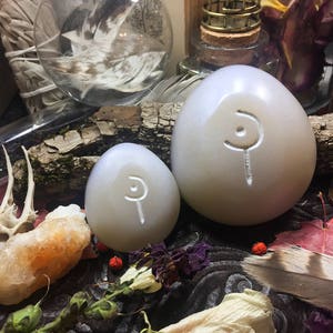 May include: Two white, egg-shaped stones with a carved symbol on each. The stones are sitting on a wooden surface with other decorative items.